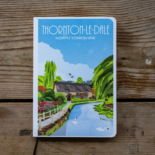 Notebook with a scenic illustration of Thorington-le-Dale, North Yorkshire, on a wooden surface.